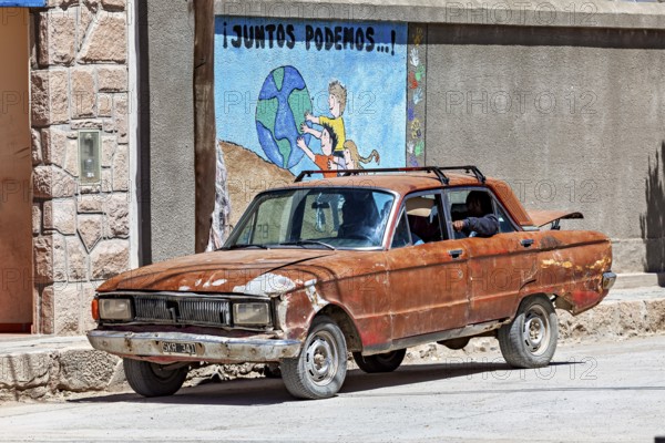 A rusty car stands on a dusty road in front of a colorful mural, Old car in traffic in San Antonio de los Cobres in Argentina
