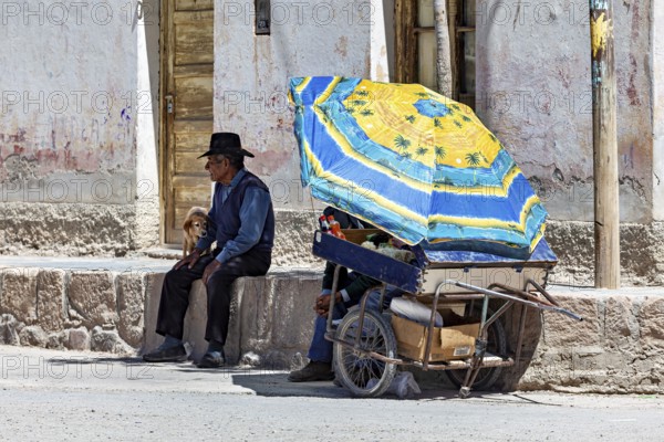 Man and child sitting under a blue parasol next to a cart, people in the streets of San Antonio de los Cobres, Salta province, Argentina