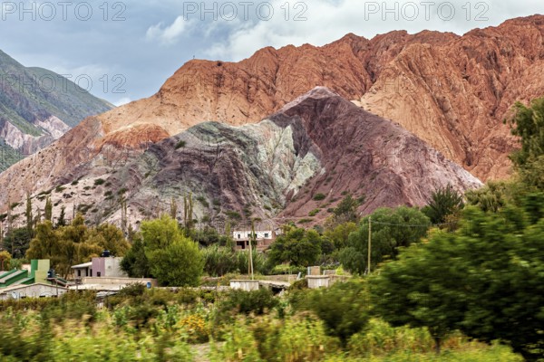 Colourful mountains rise from a green, overgrown desert under cloudy skies, the landscape and colorful rock formations of the Quebrada de Cafayate near Salta in Argentina