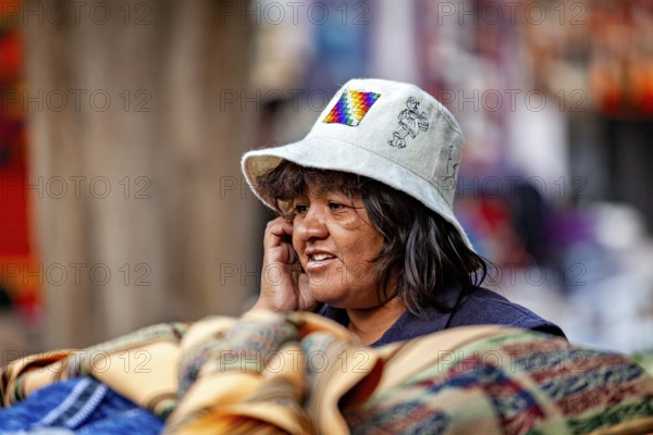 A woman wearing a traditional white hat stands next to colorful fabrics at a market, tourist and souvenir market in Santa Rosa de Tastil near Salta in Argentina