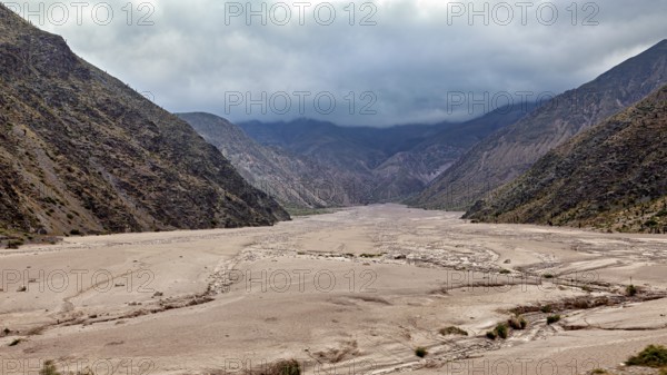 Wide valley between two dark mountain ranges under a cloudy sky, the landscape and colorful rock formations of the Quebrada de Cafayate near Salta in Argentina