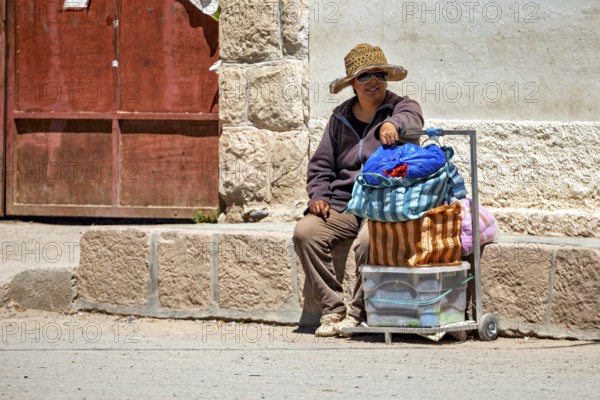 Man wearing hat sitting on the side of a road next to a loaded cart, people in the streets of San Antonio de los Cobres, Salta province, Argentina