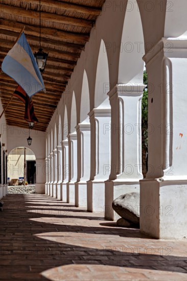 Shady arcade with white columns and wooden ceiling. An Argentinean flag is seen, The arcades of Cachi in Argentina