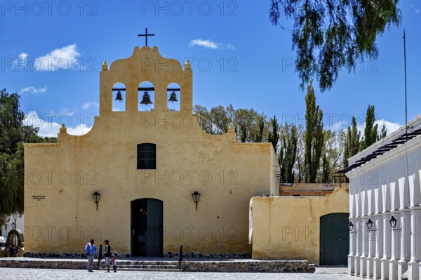Yellow church with bell tower under blue sky, people on stairs surrounded by trees, Iglesia San José de Cachi in Argentina