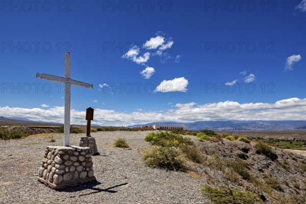 A cross stands on a hill with wide views of mountains and wide skies, roads through the countryside of the Quebrada de Cafayate near Salta in Argentina
