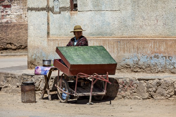Woman wearing hat sitting at a cart with a green roof on the street corner, people in the streets of San Antonio de los Cobres, Salta province, Argentina