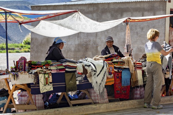 Two woman at market stalls surrounded by handmade blankets, interacting with a customer, tourist and souvenir market of Santa Rosa de Tastil near Salta in Argentina