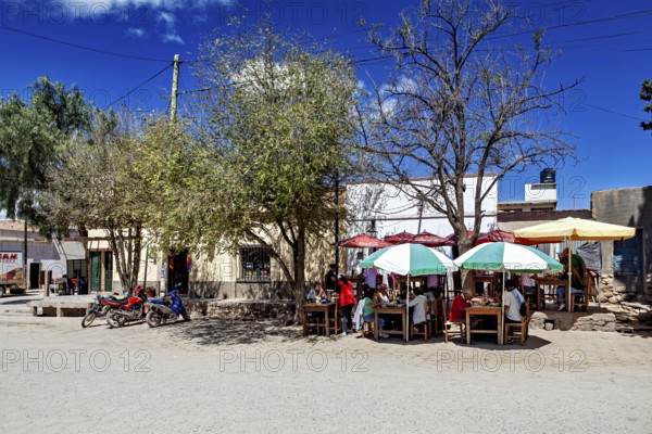 Street scene with people under colorful umbrellas at dining tables surrounded by trees and motorbikes, Downtown Cachi in Argentina