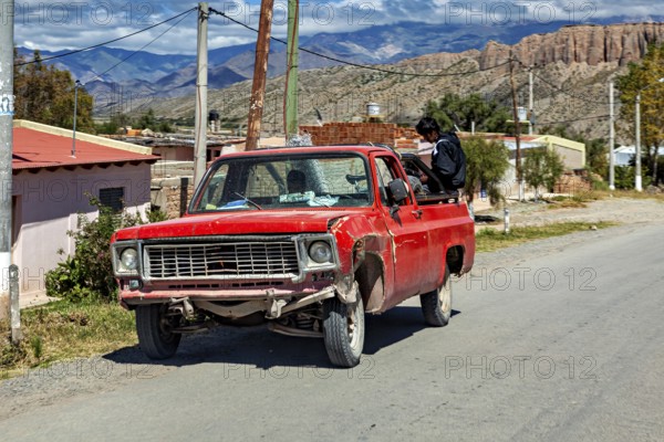 Red, slightly damaged pickup truck on a country road with a man and mountain scenery, old cars in Argentina