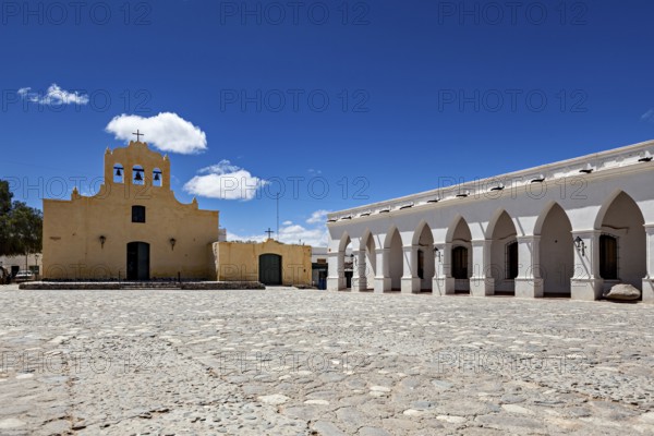 Square with yellow church and white arcades under bright blue sky, Iglesia San José de Cachi in Argentina