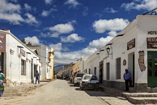 Paved street with traditional buildings, cars on the side of the road and people under a sunny sky with clouds, Downtown Cachi in Argentina