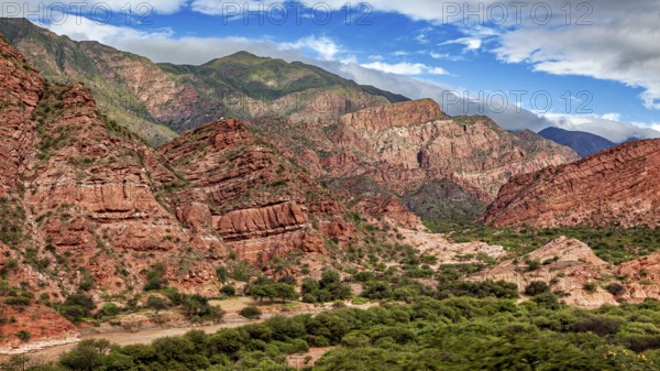 An impressive mountain landscape with red rocks, lush vegetation and a partly cloudy sky, the landscape and colorful rock formations of the Quebrada de Cafayate near Salta in Argentina