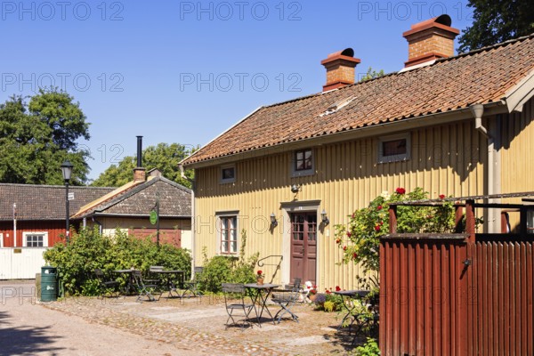 Picturesque Scandinavian courtyard featuring a traditional wooden building with garden furiture on a cobblestone patio surrounded by potted plants and blooming flowers a small town atmosphere under a bright blue sky a sunny summer day, Lidköping, Sweden