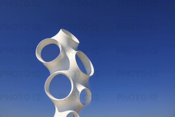Contemporary white abstract sculpture composed of smooth tubular forms with large circular openings against a deep blue sky. Geometric shapes at a modern artworks in public spaces, Lidköping, Sweden