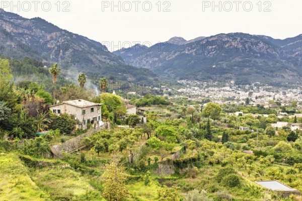 Serene rural landscape featuring a traditional stone house surrounded by vibrant greenery and terraced gardens with a city in the mountain valley, Sóller, Mallorca, Spain