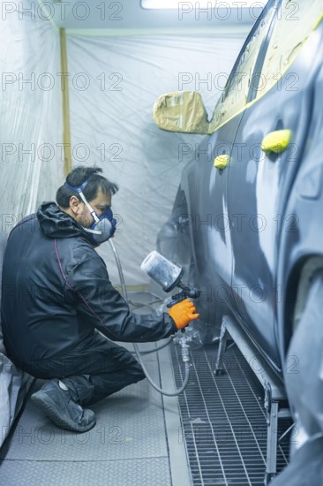Auto body technician wearing mask and gloves sprays primer onto a car door panel inside a ventilated paint booth, preparing the surface for professional refinishing