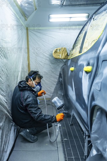 Mechanic in respirator and gloves crouching in a paint booth, spraying a sedan's side panel with a spray gun, focused on precise automotive repainting and repair