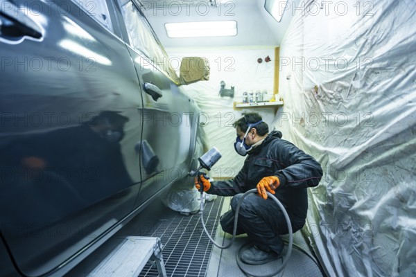 Mechanic wearing respirator mask and orange gloves spraying dark car in a professional paint booth, performing auto body repair and re painting services