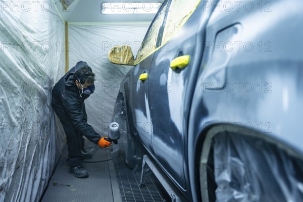 Auto body technician working in a repair shop, carefully applying paint primer to a vehicle door and fender with a spray gun inside a protective spray booth