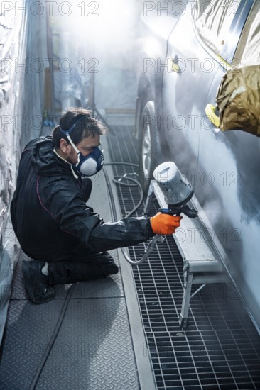 Male auto body mechanic in protective mask and suit applying a fresh coat of gray paint to a car door with a spray gun in a dusty professional paint booth