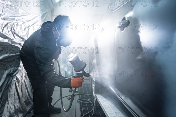 Auto mechanic applying primer paint to a car body panel using a spray gun in a well lit automotive workshop paint booth for vehicle repair and maintenance