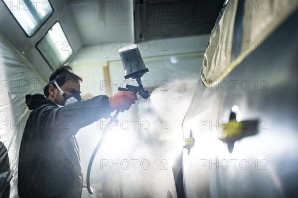 Auto body technician wearing protective mask and gloves applying a fresh coat of paint primer to a car panel with a spray gun in a professional automotive repair shop for restoration