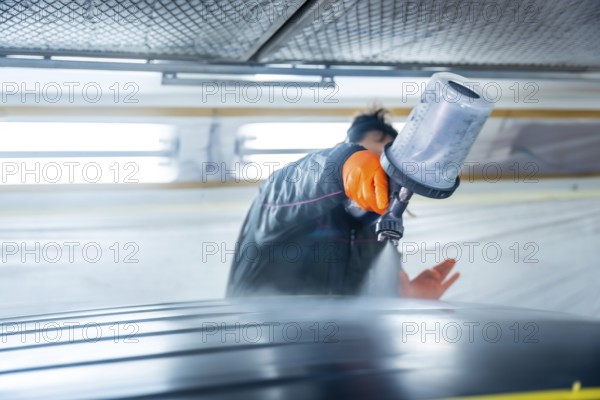 Auto body technician wearing protective gloves painting a vehicle car roof using a spray gun in a well lit automotive service workshop, ensuring high quality refinishing and repair work