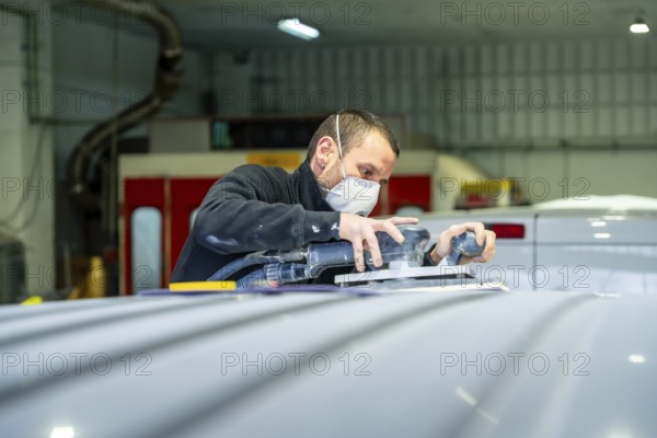 Auto body technician wearing protective mask sanding a vehicle roof with an orbital sander at a professional car repair and paint shop, performing automotive refinishing work