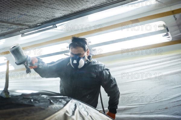 Auto body technician working in a specialized workshop, wearing a respirator mask and protective suit while applying fresh paint to a car part with a paint gun