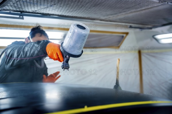 Automotive technician wearing safety gloves and mask meticulously applying a fresh coat of paint to a car roof using a professional spray gun in a clean paint booth