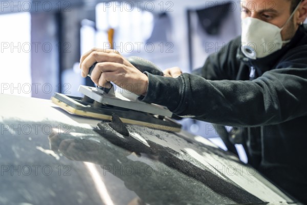 Professional mechanic performing essential work in a car body and paint shop, sanding the vehicle's surface to prepare it for painting or finish while wearing a respirator for safety