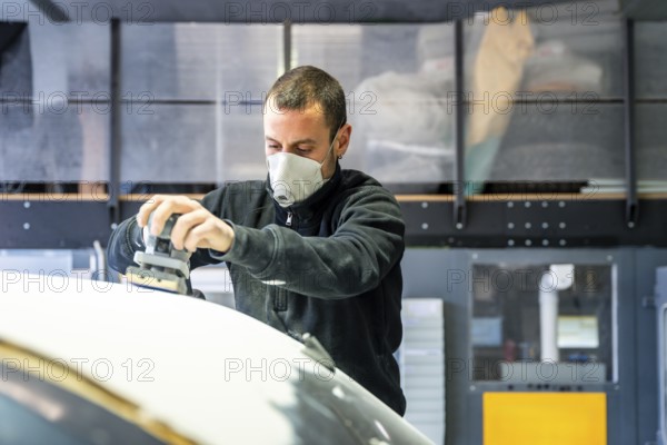 Auto body technician wearing a respirator mask using an orbital sander to prepare a car surface for painting, managing dust in a professional garage environment