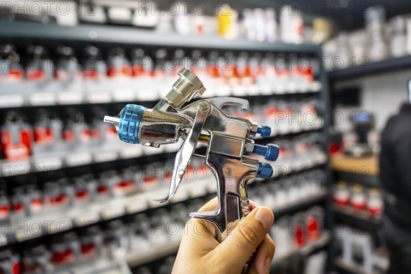 Hand holding a high pressure spray gun, selecting paint colors from shelves in an automotive refinishing workshop, preparing for car body repair and painting