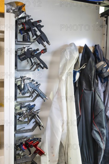 Modern spray guns and protective gear, including a disposable suit and a respirator mask, hanging in a car body paint shop, ready for vehicle repair and refinishing work