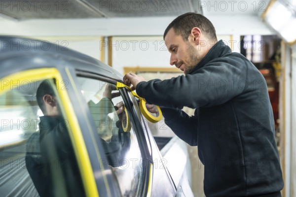 Automotive technician carefully applying yellow masking tape around a car window and trim before a paint job in a professional auto body repair workshop