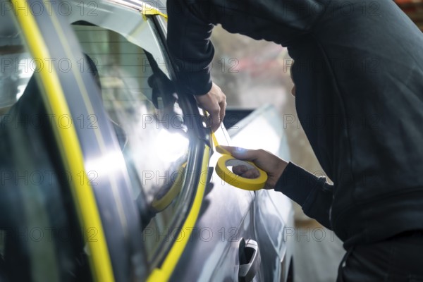 Auto body technician hands applying yellow masking tape around car window trim in a workshop, preparing the vehicle for precise paint work and restoration with care and focus