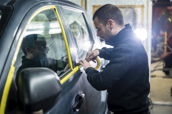 Auto body shop worker carefully applying yellow masking tape to a car door window, preparing the vehicle for a new paint job in a professional automotive repair service
