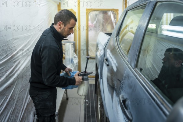 Worker preparing car body for painting in an automotive repair facility, applying solution to the vehicle's surface for final sanding and surface preparation before the paint application process