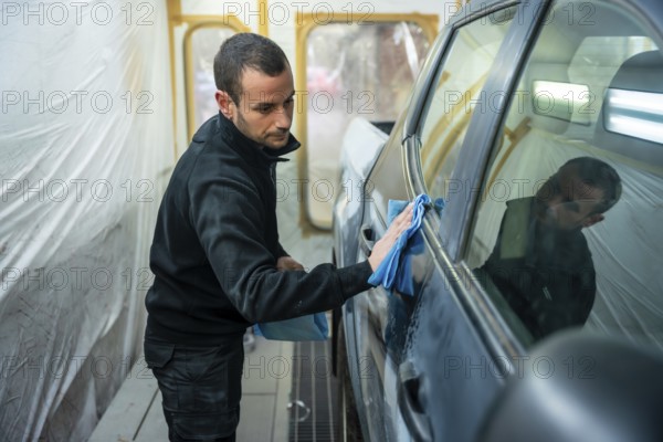 Man carefully wiping car body panels with a blue microfiber cloth inside a professional spray booth, preparing the vehicle for painting precise automotive detailing and repair work