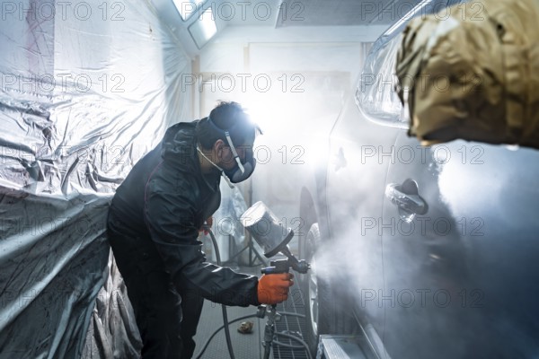 Professional automotive technician applying a fresh coat of paint to a vehicle in a specialized spray booth, wearing a respirator mask and protective gear during the careful repainting process