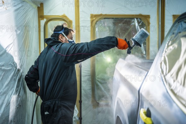 Auto body painter wearing protective equipment and holding a spray gun, expertly applying a fresh coat of paint to a vehicle door during the refinishing process inside a dedicated paint booth