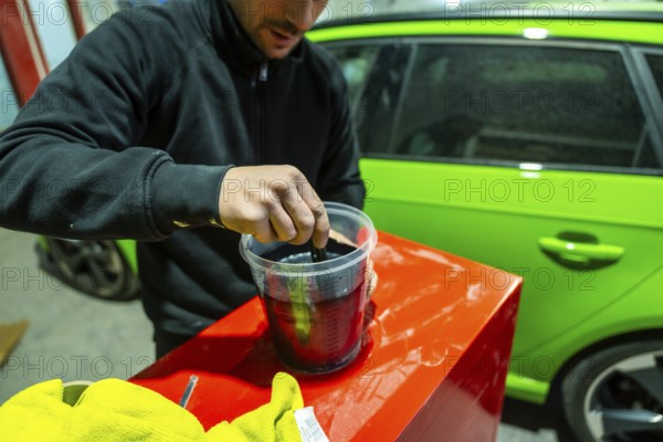 Male worker preparing automotive paint in a plastic container with a lime green vehicle visible in the background, focusing on the careful process of color matching and repair