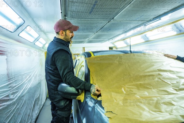 Skilled technician carefully masking vehicle with brown paper and tape, preparing it for a professional automotive paint job in a well lit car repair shop