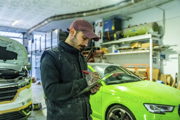 Auto body technician wearing a cap and overalls writing on a checklist, inspecting vehicles in a professional garage during a car repair and painting process