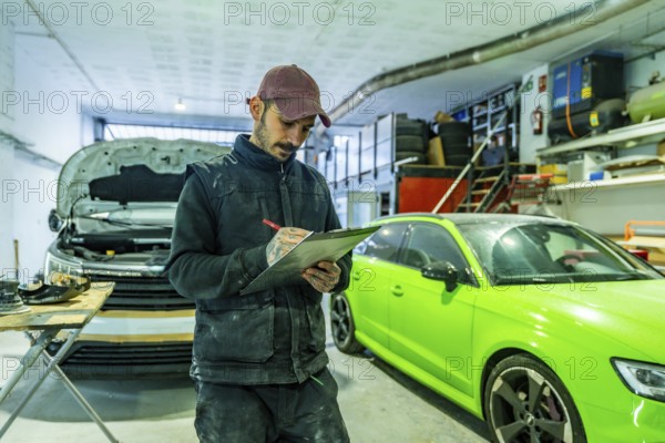 Mechanic with tattooed arms checks inspection checklist on a clipboard in a busy auto body and paint shop, repaired cars and tools visible in the background