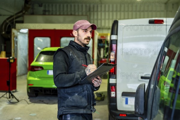 Auto body shop technician checking details on a white van and a green car in the background, working with a clipboard and pen in a professional automotive service garage