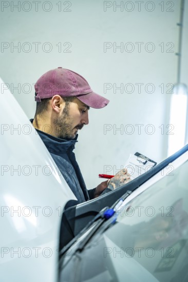 Auto technician wearing a cap writing details on a clipboard while inspecting vehicle condition in a professional garage or body shop, ensuring quality service and maintenance