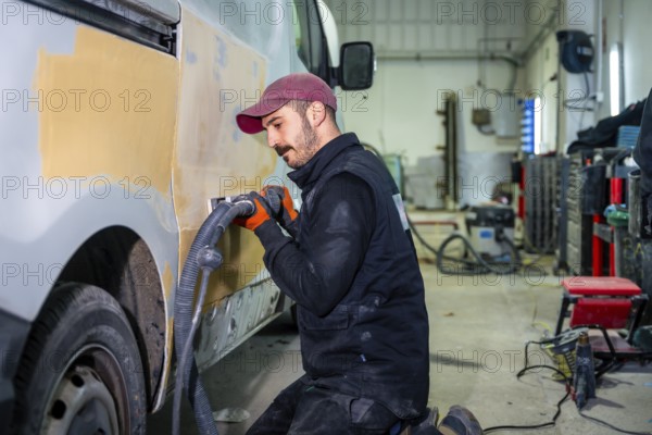 Auto body technician working on a van, utilizing a power sander with vacuum attachment to smooth filler on the vehicle's side panel at an automotive repair workshop