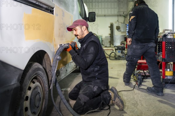 Male auto body worker kneeling and using a power sander with vacuum attachment on a van door, preparing the surface for painting in a professional repair shop alongside another technician