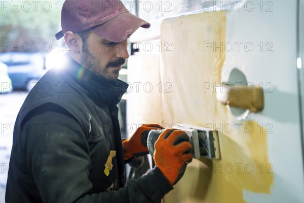 Skilled auto body worker wearing work gloves and a cap carefully sanding a car door with filler applied, preparing the surface for painting during vehicle repair in a workshop